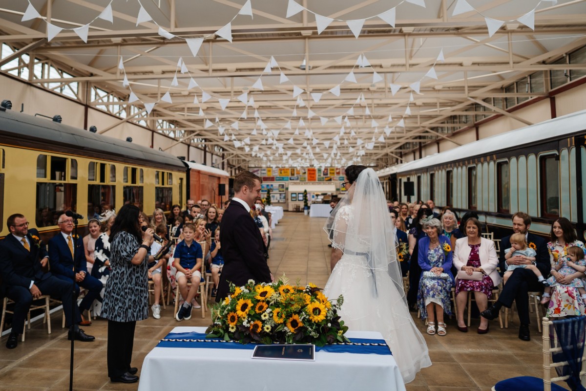 Liz and Paul’s wedding ceremony taking place inside a vintage railway venue with guests seated around them.