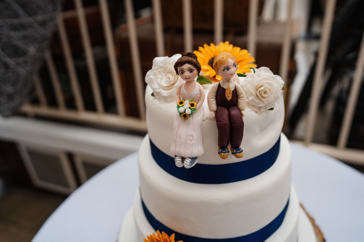 Wedding cake decorated with miniature bride and groom figures, white flowers and a blue ribbon.