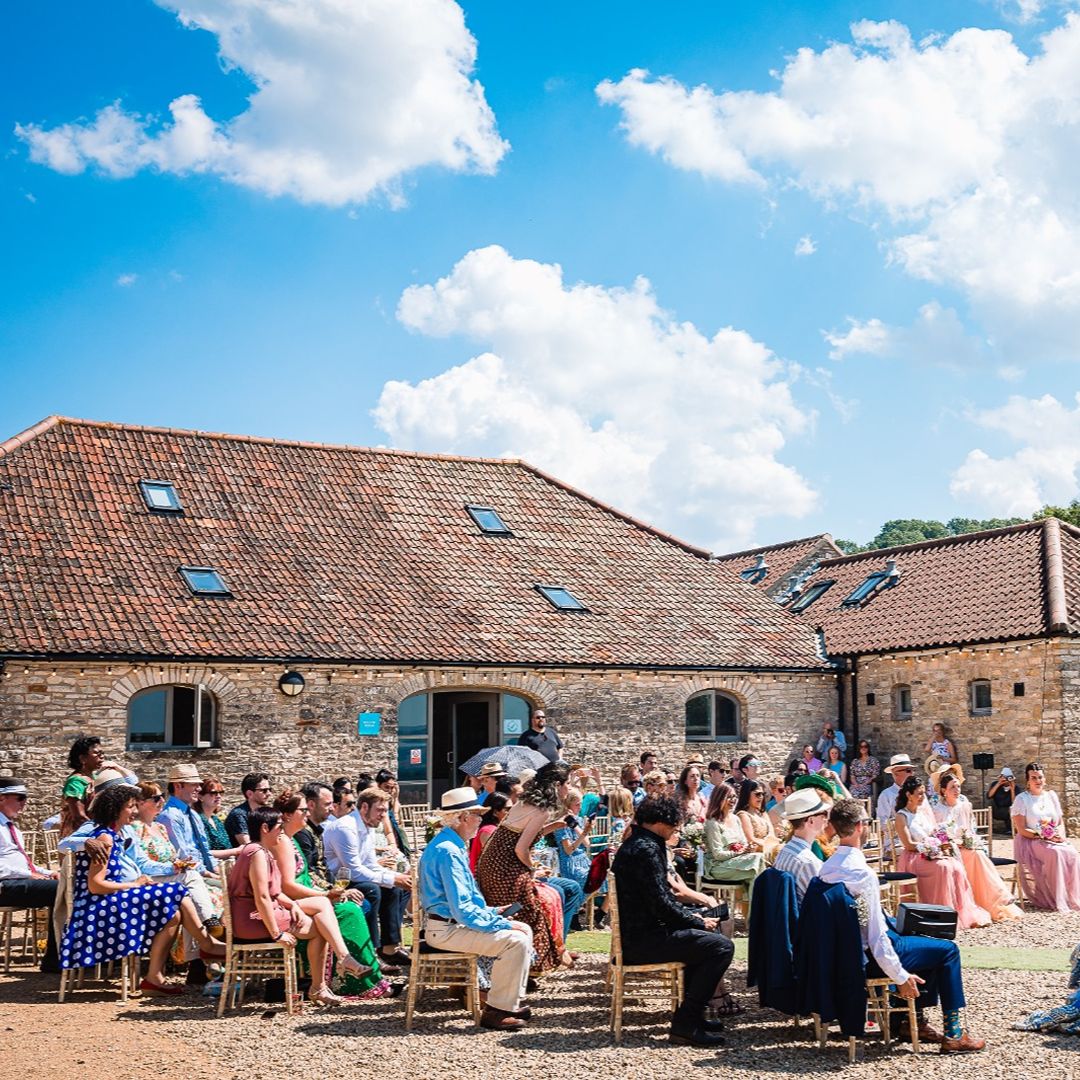 Wedding guests seated outdoors in a courtyard, gathered for a ceremony at a rustic stone venue beneath a bright blue sky.