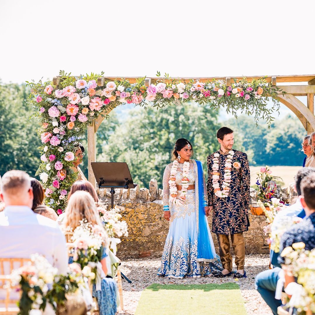 Couple standing beneath a floral wedding arch during an outdoor ceremony, surrounded by seated wedding guests.
