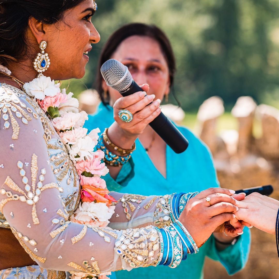 Bride in traditional blue and silver wedding attire holding a microphone while touching a guest’s hand during an outdoor ceremony.