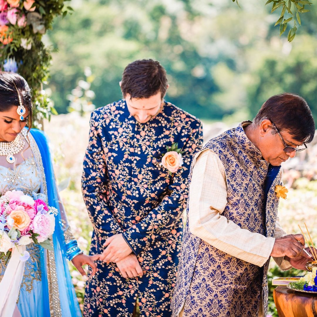 Couple in traditional wedding attire standing beside a family member during an outdoor wedding ceremony.