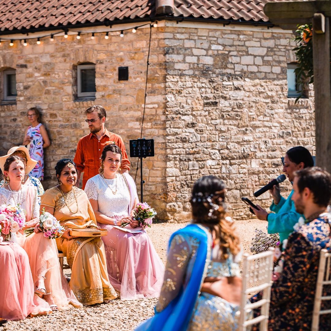 Wedding guests seated outdoors as a celebrant speaks during a ceremony, with the bride and groom visible in the foreground.