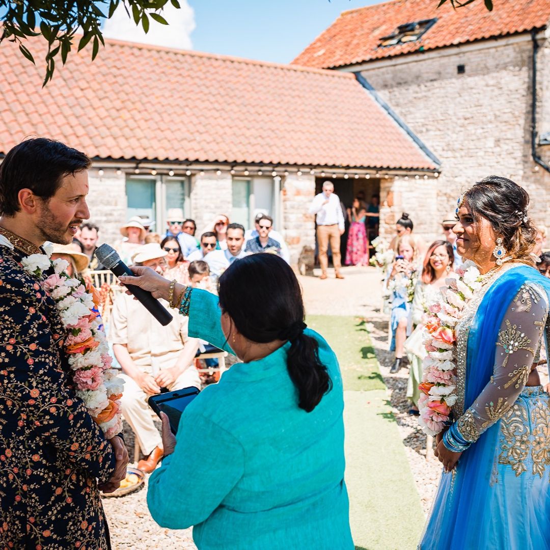 Couple in wedding attire standing outdoors as a celebrant speaks into a microphone during their wedding ceremony, with guests seated nearby.