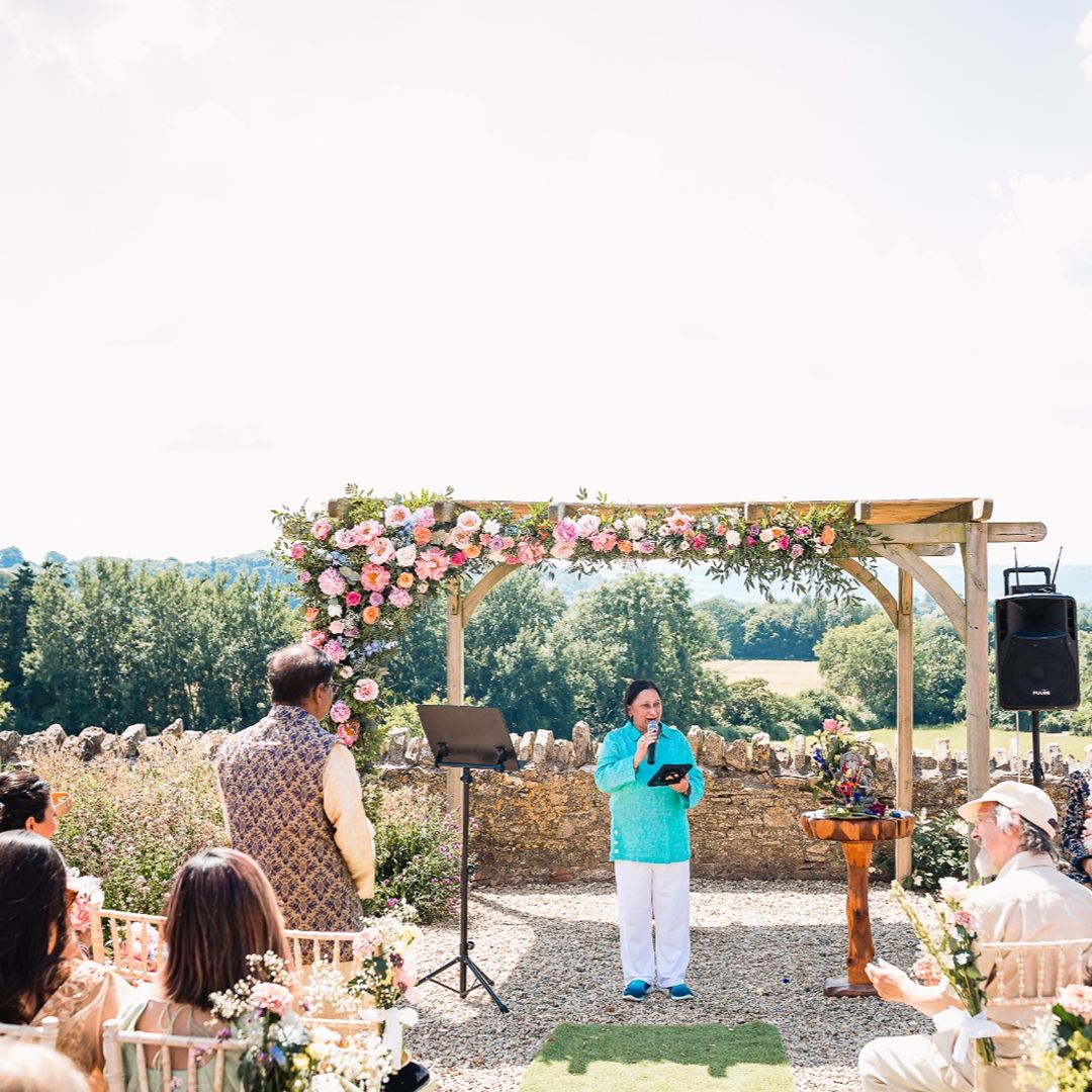 Outdoor wedding ceremony with a celebrant standing beneath a floral-decorated wooden arch, surrounded by seated guests in a countryside setting.