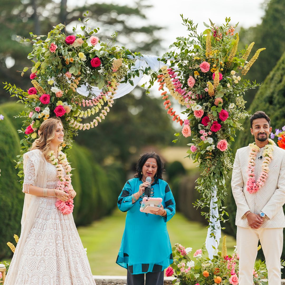 Natasha and Aaron stand beneath a floral wedding arch with their celebrant, Sonal Dave during their multicultural outdoor wedding ceremony.